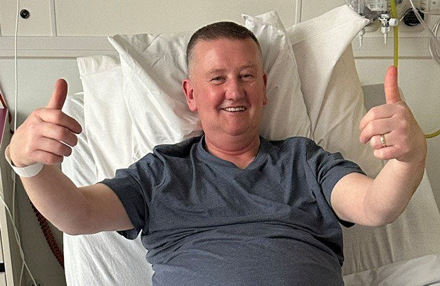 Adrian Sharkey sitting up in bed in a grey T-shirt, smiling and giving two thumbs up.