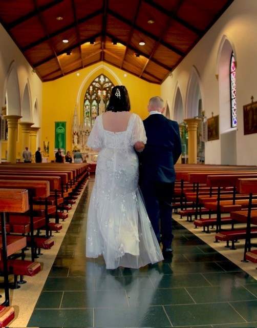 Adrian, in a dark suit, walking his daughter Lauren in her wedding dress down the aisle of a church, seen from behind.